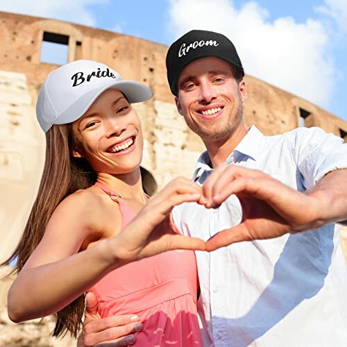 Happy couple in bride and groom caps making a heart with hands.