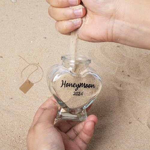 Hands pouring sand into a heart-shaped bottle labeled 'Honeymoon 2024'.